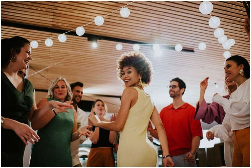 A group of eight people are gathered together indoors, smiling and dancing under white string lights in a joyful, candid setting.