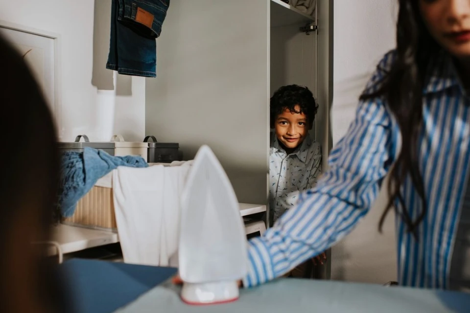 A woman does ironing, with a child looking out from inside the closet. Domestic scene with laundry and household objects visible.