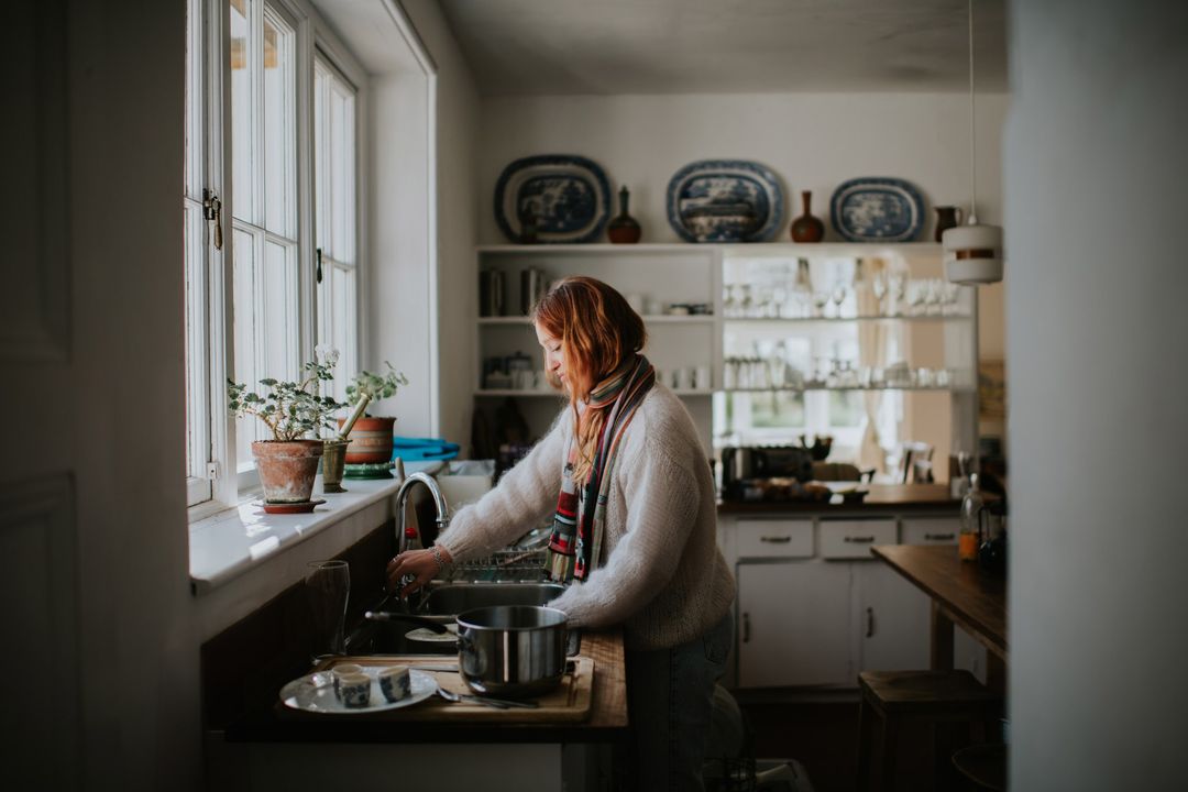Woman stands at a sink in a domestic kitchen with a large window, plants, plates, and dishware. She is wearing a striped scarf.