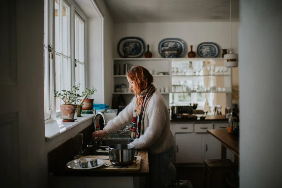 Woman stands at a sink in a domestic kitchen with a large window, plants, plates, and dishware. She is wearing a striped scarf.