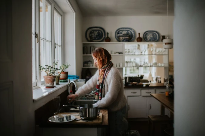 Woman stands at a sink in a domestic kitchen with a large window, plants, plates, and dishware. She is wearing a striped scarf.