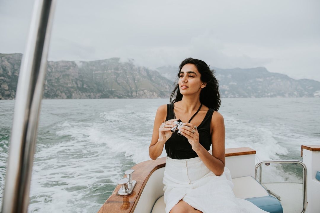 Young woman enjoys a boat ride on a cloudy day, holding binoculars and gazing at distant mountains over the water.