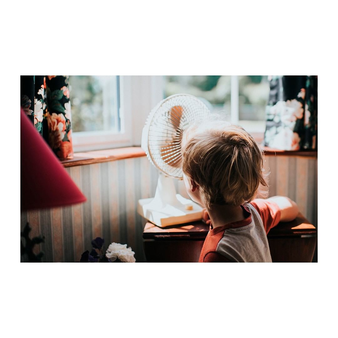 A young child with blonde hair stands in front of a white fan on a wooden table, bathed in sunlight near a window with floral curtains.