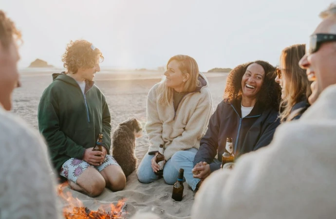 Group of friends and a dog gather around a beach bonfire at sunset, enjoying beers and each other's company in a candid, relaxed setting.