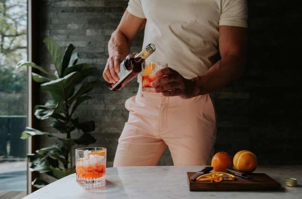 Person pours cocktail into glass with ice and orange peel; second glass and oranges on marble table with neutral wall and plant.