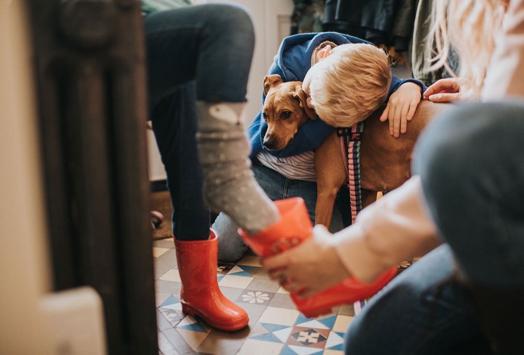 Blond child in blue hugs brown dog; red rain boots on patterned floor; partial views of two other people visible.