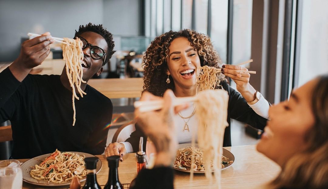 Three smiling people enjoy eating noodles with chopsticks at a restaurant, with bowls of noodles and sauces on the wooden table.