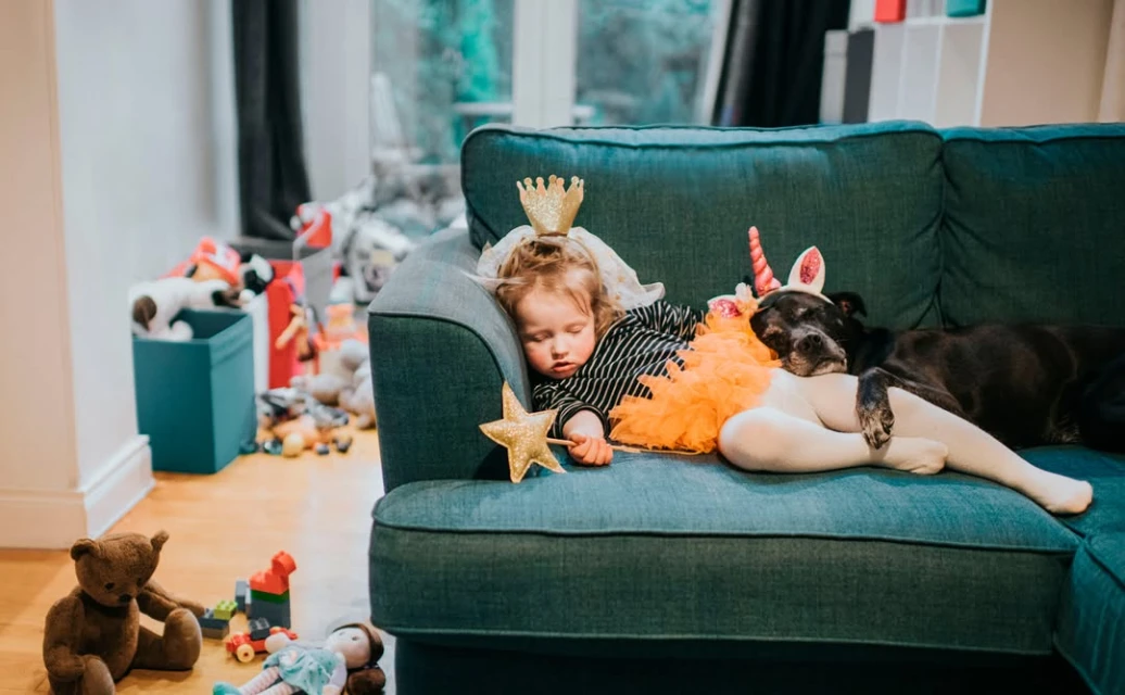 A toddler and a black dog sleeping together on a green couch, both wearing costume headwear, in a bright, messy living room.
