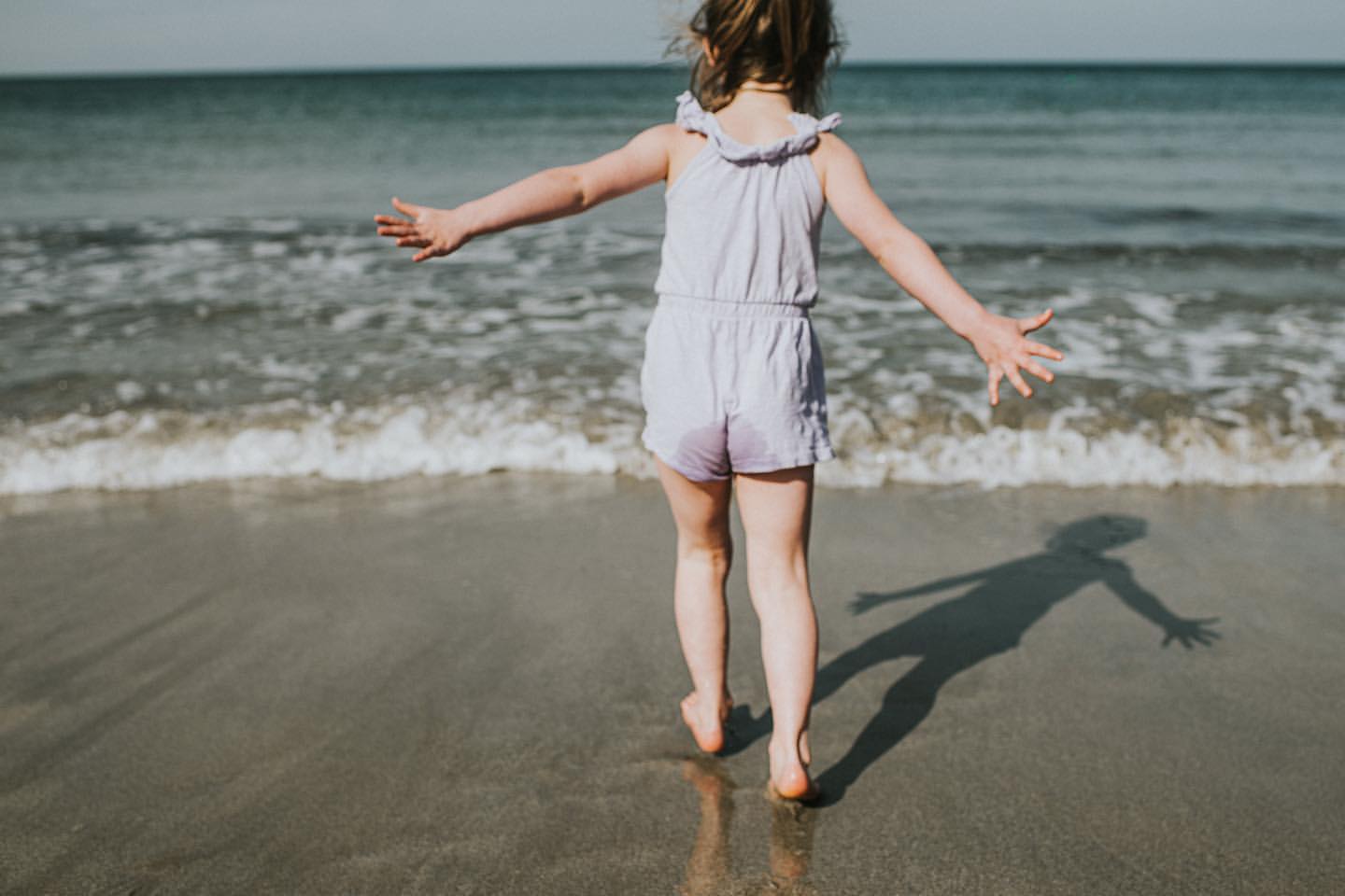 Child in purple playsuit standing on a sandy beach with arms outstretched towards the ocean waves, enjoying a sunny day at the seaside.