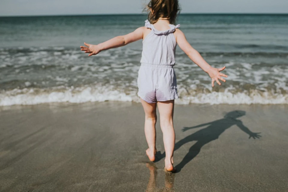 Child in purple playsuit standing on a sandy beach with arms outstretched towards the ocean waves, enjoying a sunny day at the seaside.