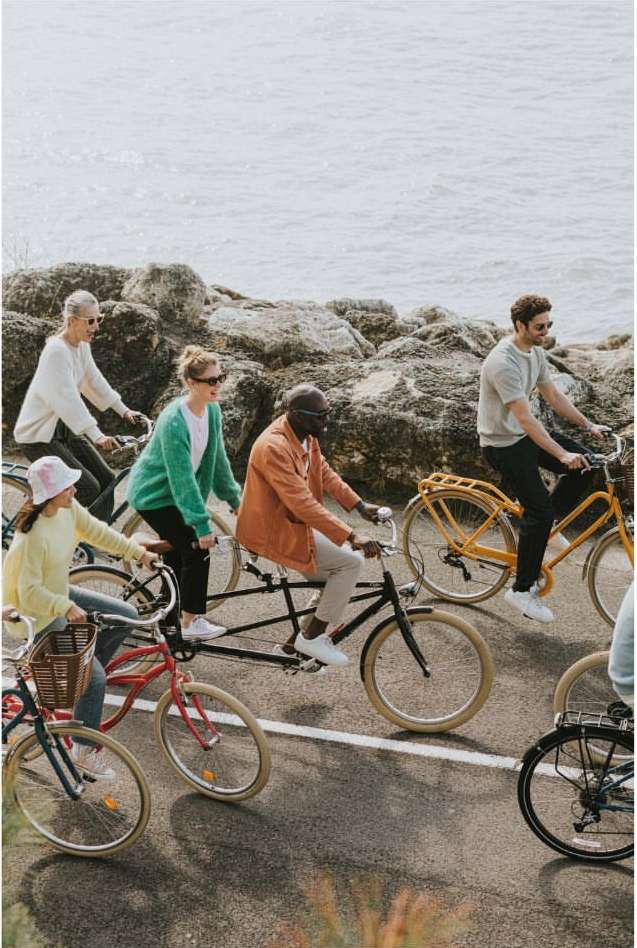 Group of diverse people riding bikes on paved road near rocky coastline, wearing casual clothing, enjoying an active lifestyle in daylight.
