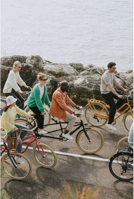 Group of diverse people riding bikes on paved road near rocky coastline, wearing casual clothing, enjoying an active lifestyle in daylight.