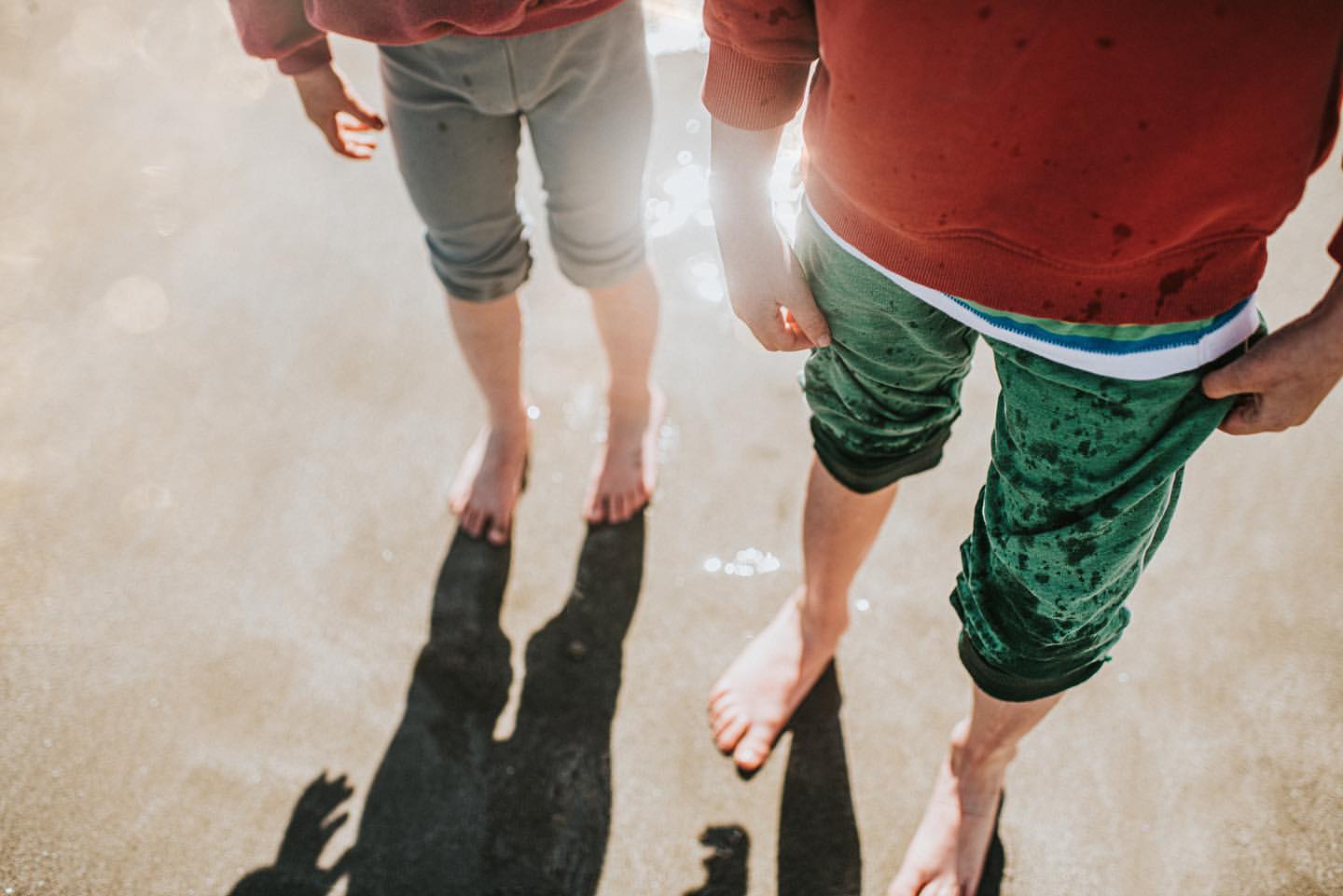 Two children are standing barefoot in shallow water on a sunny day, wearing shorts and tops, with long shadows cast on the beach.