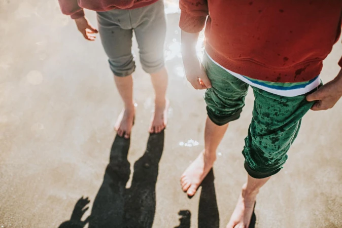 Two children are standing barefoot in shallow water on a sunny day, wearing shorts and tops, with long shadows cast on the beach.