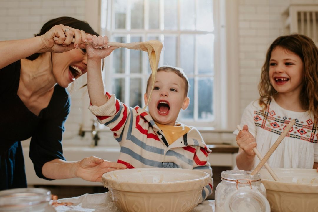 Mother and two kids joyfully playing with dough in a bright kitchen, wearing casual clothes, creating a warm, family moment.