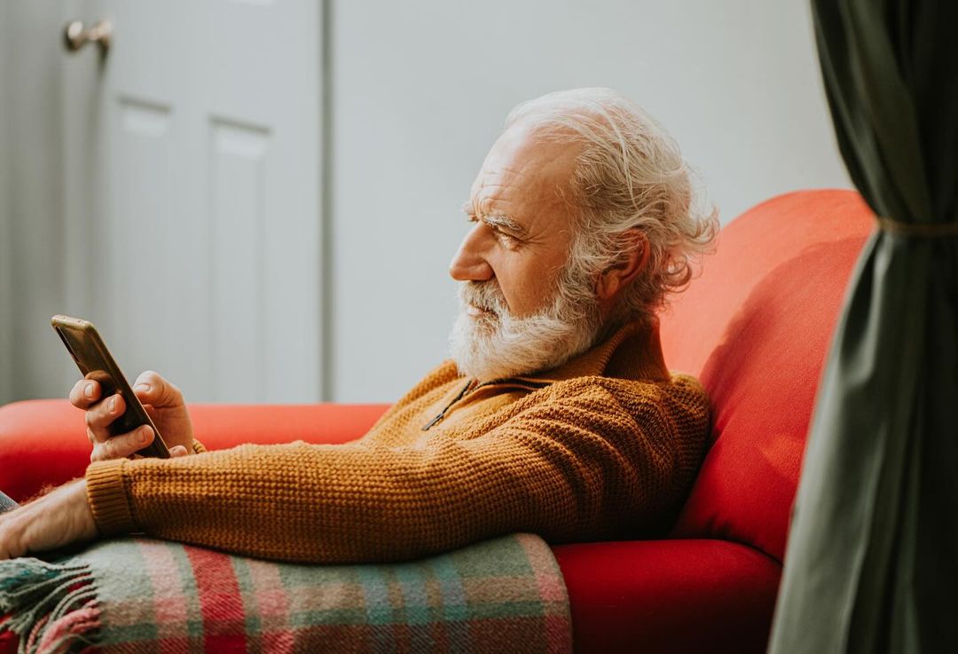 Elderly man with a white beard sits on a red couch using his phone, wrapped in a blanket, in an indoor setting with natural lighting.