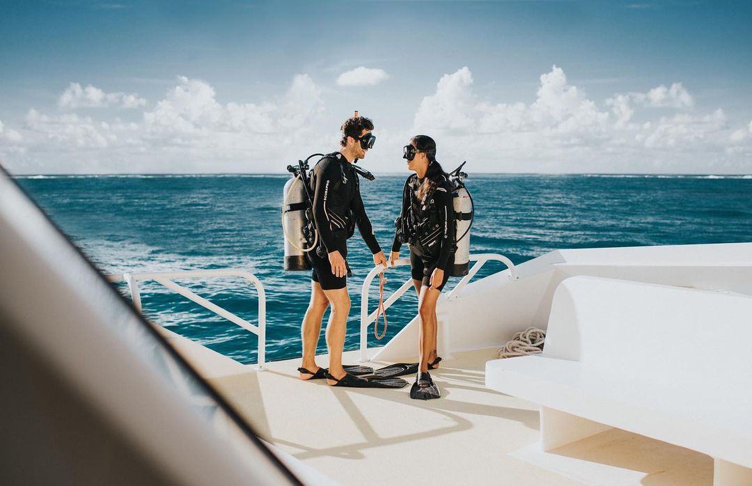 Two scuba divers in wetsuits on a boat deck with tanks, masks, and fins, standing near the ocean with a cloudy sky in the background.