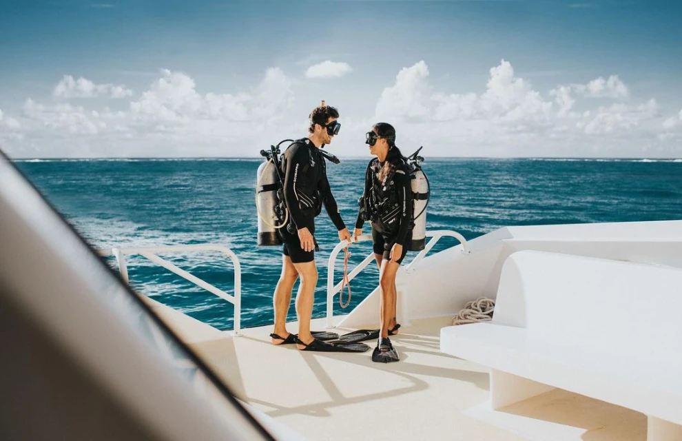 Two scuba divers in wetsuits on a boat deck with tanks, masks, and fins, standing near the ocean with a cloudy sky in the background.