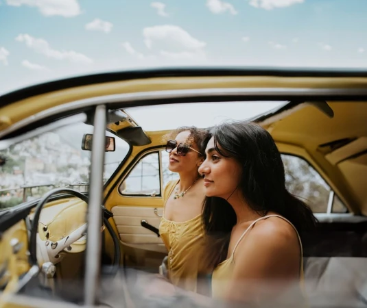 Two women in a yellow convertible car, smiling and looking away from camera, enjoying a sunny day outdoors.