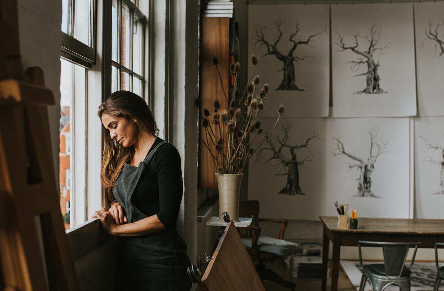 Woman in apron looks out a window in an art studio with illustrations of trees on the wall and art supplies on a wooden table.