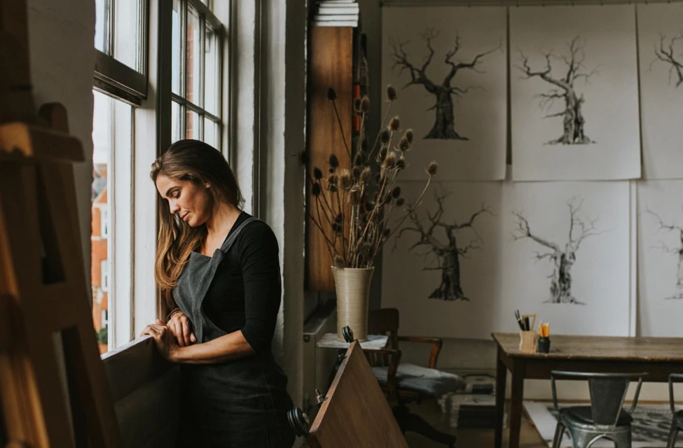 Woman in apron looks out a window in an art studio with illustrations of trees on the wall and art supplies on a wooden table.
