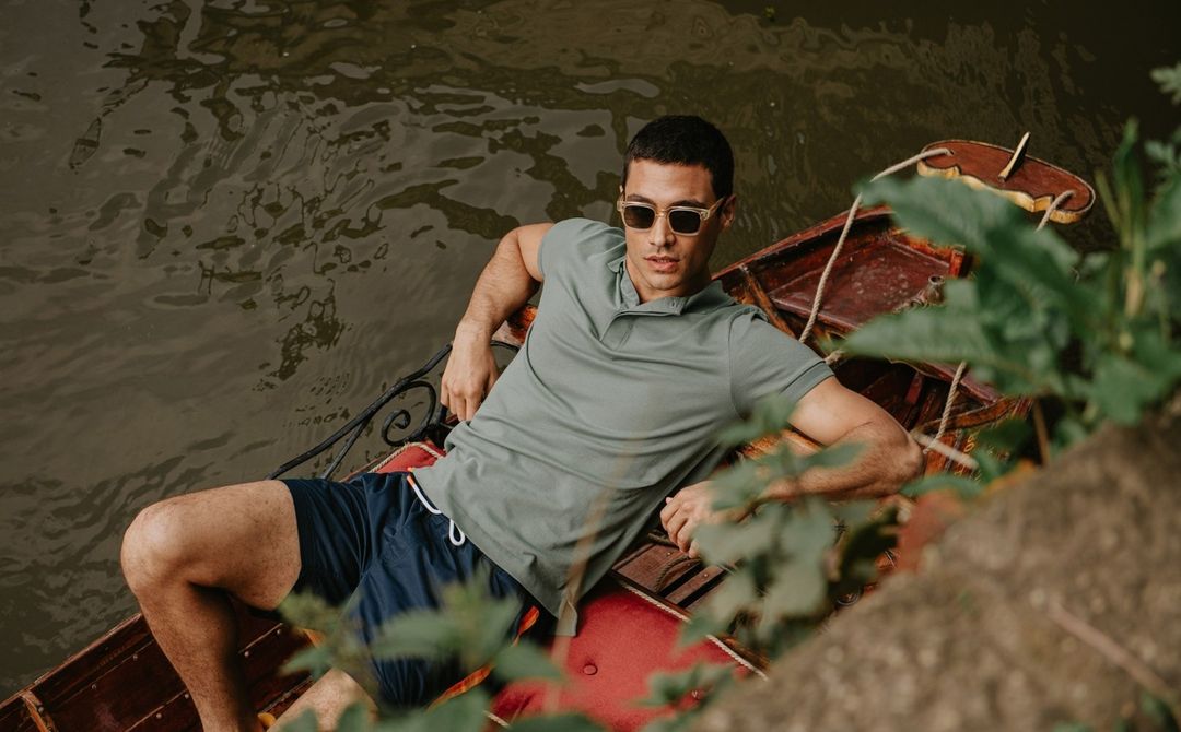 A man wearing sunglasses reclines in a small boat on the water, wearing a polo shirt and shorts in outdoor setting.