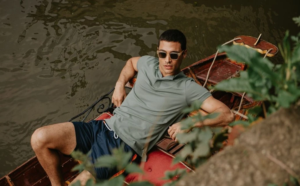 A man wearing sunglasses reclines in a small boat on the water, wearing a polo shirt and shorts in outdoor setting.