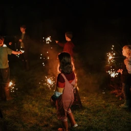 Children holding sparklers outdoors at night, illuminating the dark grassy garden with bright orange sparks and warm glowing light.