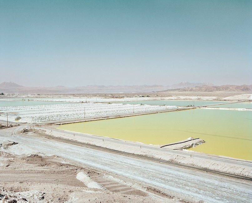 Expansive view of a desert landscape featuring colorful salt ponds, dry terrain, and distant mountains under a clear, light-blue sky.