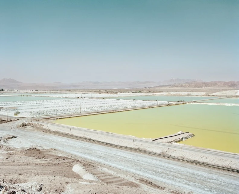 Expansive view of a desert landscape featuring colorful salt ponds, dry terrain, and distant mountains under a clear, light-blue sky.