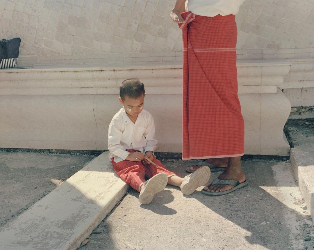 Young boy sits with an adult in a bright white and red traditional setting, bathed in the sun's light with visible shadows.