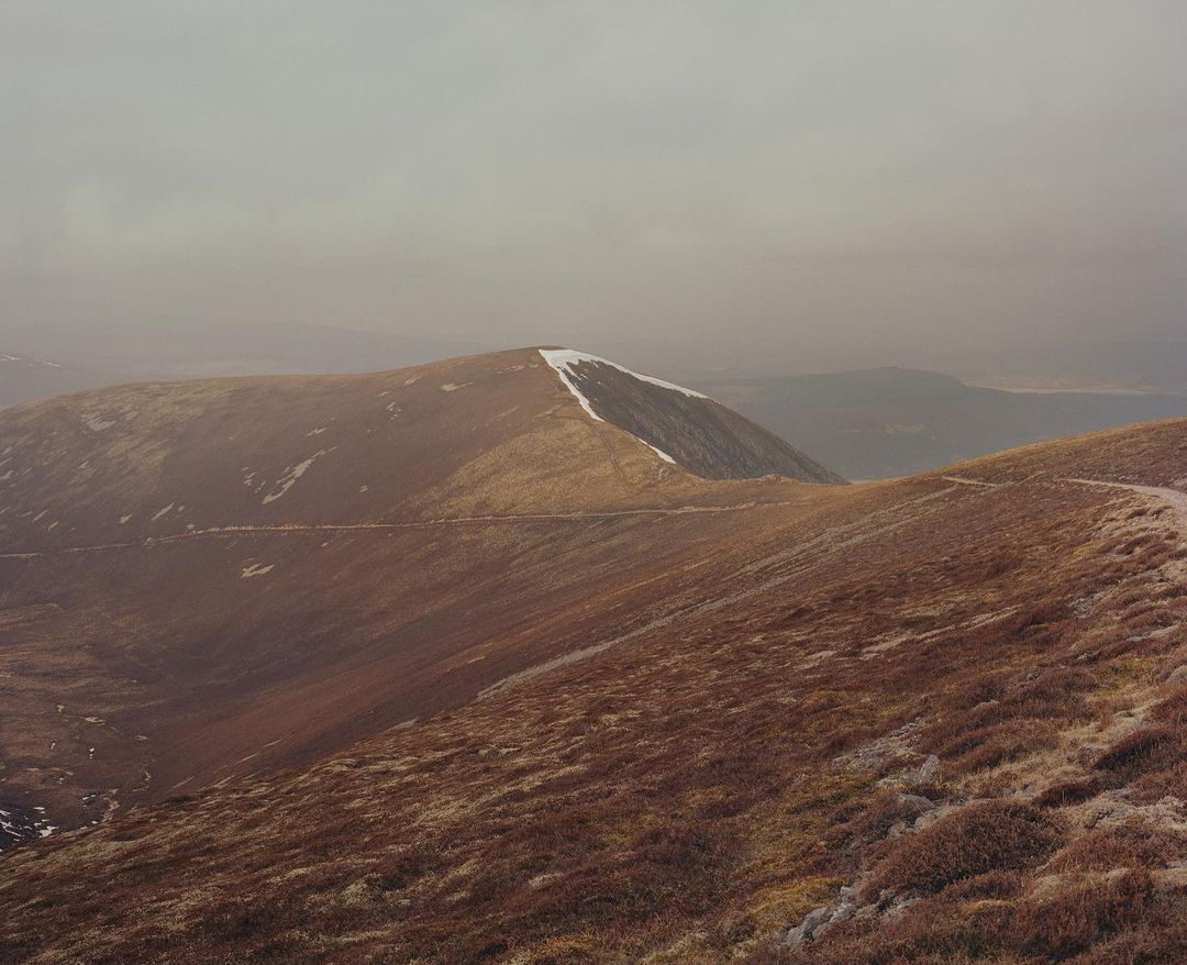 Wide shot of a rural mountain landscape with a dirt trail, snow-capped peaks, and overcast sky, creating a tranquil and natural scene.