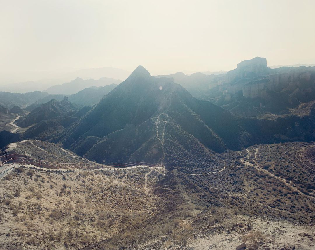 Scenic landscape featuring a mountainous region with rugged peaks, winding paths, and arid vegetation, under an overcast sky, capturing natural beauty.