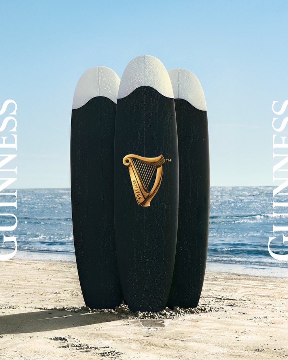 Three black surfboards with white tops stand on a sandy beach with the Guinness logo displayed on the center surfboard.