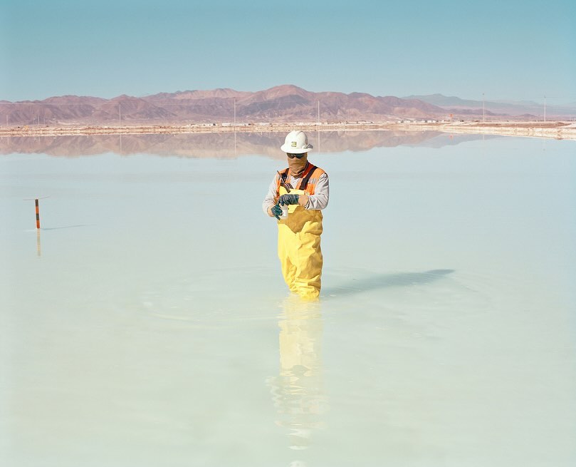 A worker wearing a hardhat and yellow waterproof overalls stands in shallow water with mountains and blue sky in the background.