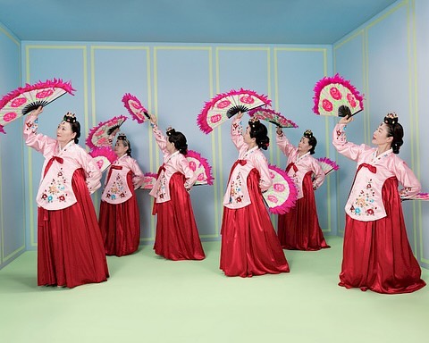 Seven Korean dancers in traditional pink and red hanboks gracefully perform with colorful fans in a pastel-colored indoor setting.