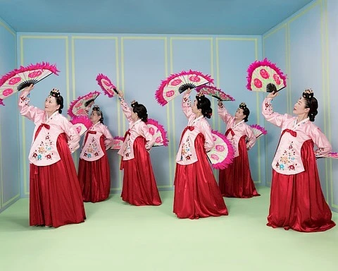 Seven Korean dancers in traditional pink and red hanboks gracefully perform with colorful fans in a pastel-colored indoor setting.