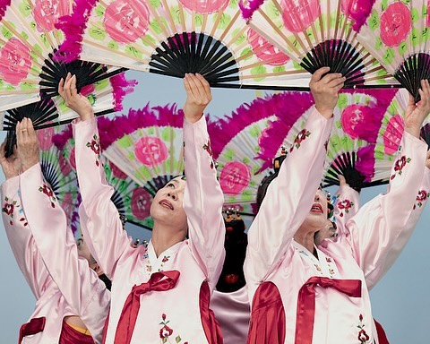 Group of women in traditional Korean dresses hold fans in a choreographed dance performance, set against a bright blue sky.