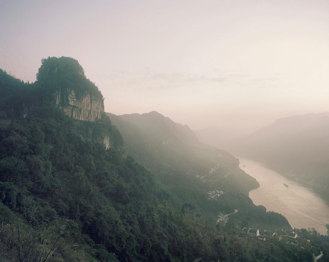 Scenic mountain landscape with lush vegetation, a winding river, and a village nestled in the valley, all under a soft, hazy sky.