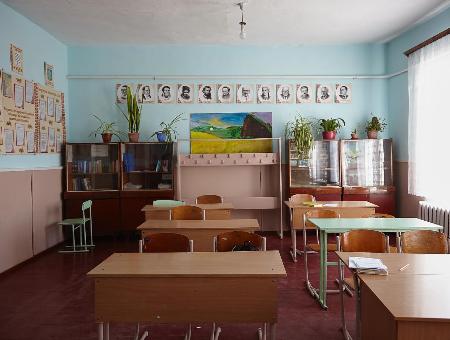 Classroom interior featuring rows of wooden student desks and chairs, bookshelves, and educational posters in a school setting.