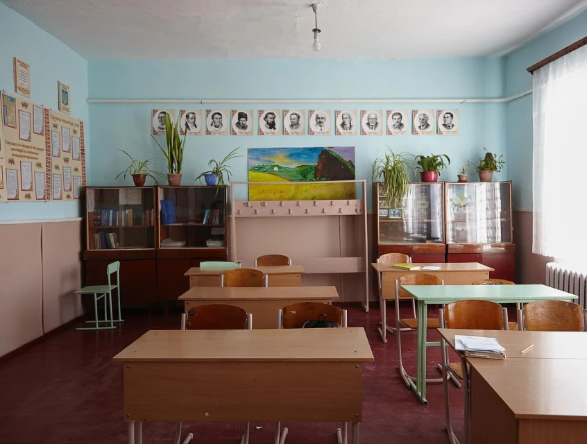 Classroom interior featuring rows of wooden student desks and chairs, bookshelves, and educational posters in a school setting.