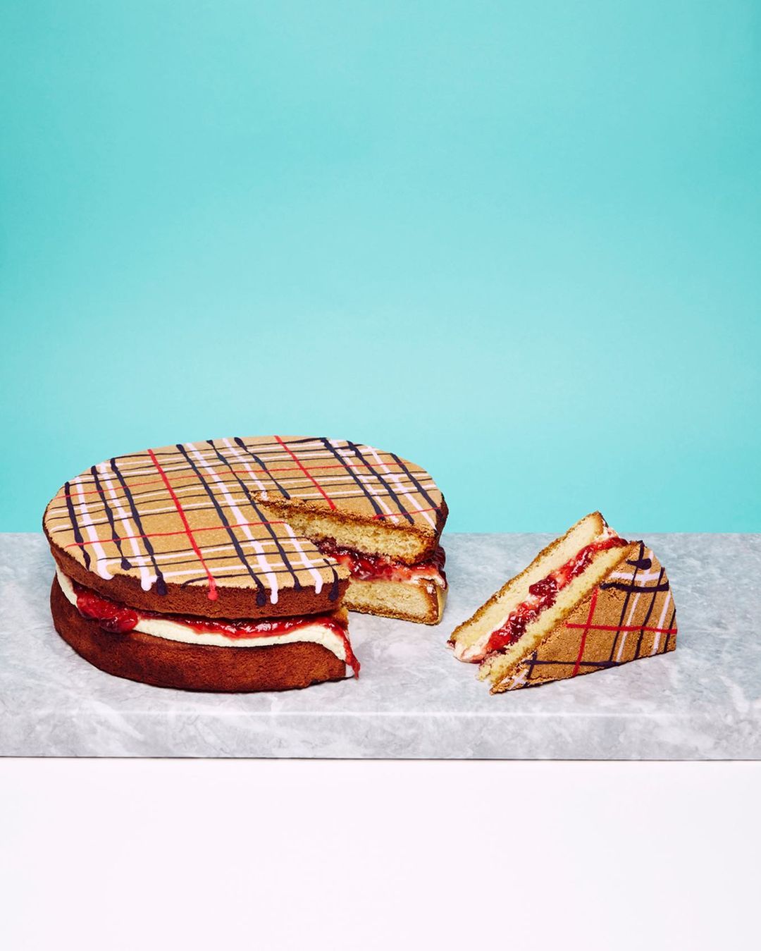 Tiered cake with plaid pattern and strawberry filling, displayed on a marble surface against a teal background in a studio setting.
