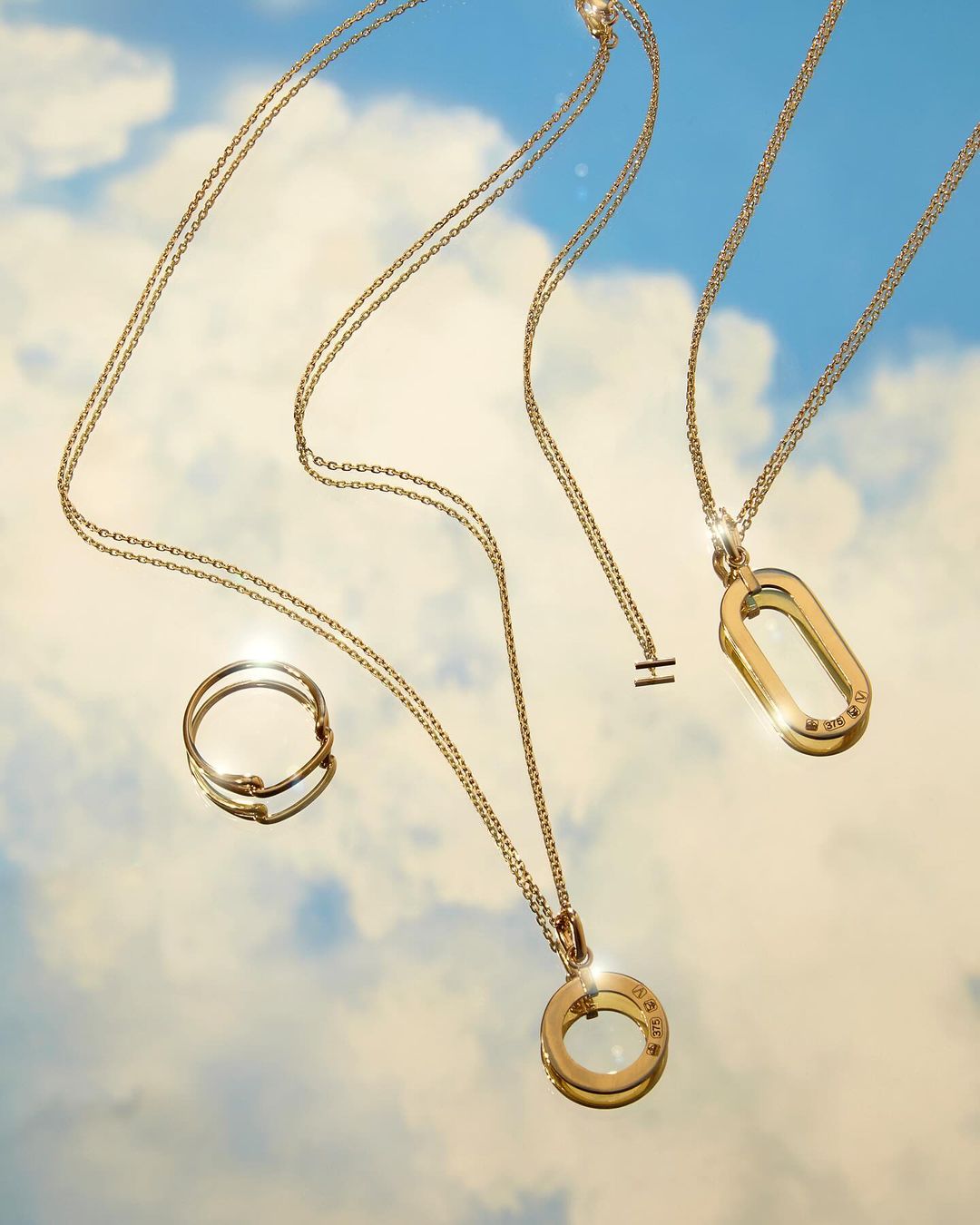 Elegant jewelry display featuring gold necklaces and a ring against a serene blue sky with soft, scattered clouds creating a dreamy backdrop.