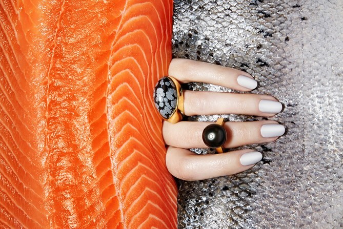 Close-up product shot of a manicured hand adorned with rings, placed against a backdrop of salmon and shimmering fish scales.