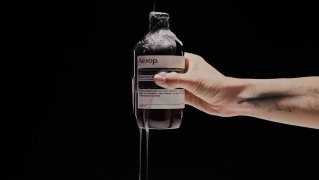 Hand holding a bottle of Aesop product with liquid pouring over it on a black background, showcasing beauty and skincare aesthetics.