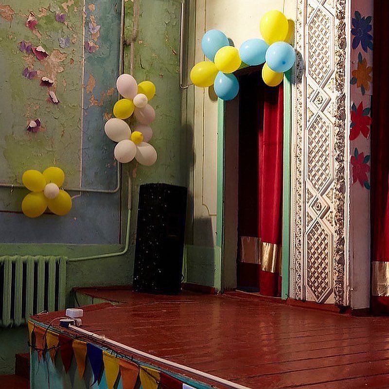 Festive stage setting with balloon decorations in blue, yellow, and white, complemented by colorful bunting and vintage architectural details.