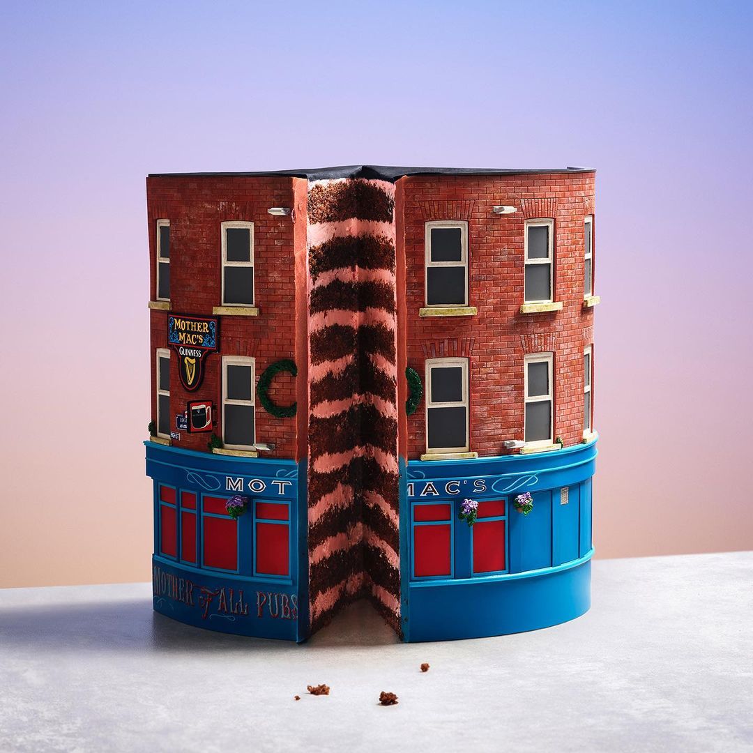 Unique cake shaped like a corner pub, featuring brick facade, blue trim, and detailed signage. It has a slice cut to show the cake layers.
