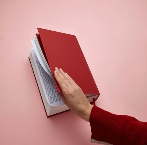 Close-up of a hand turning the page of a red hardcover book with white pages, set against a light pink background.