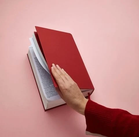 Close-up of a hand turning the page of a red hardcover book with white pages, set against a light pink background.