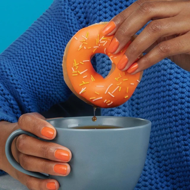 A person dips an orange frosted donut with white and orange sprinkles into a grey mug in front of a blue background.